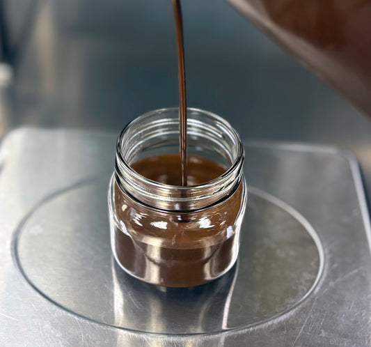 A small clear glass jar sits on a metal scale while a thick brown liquid is being poured into it from above.