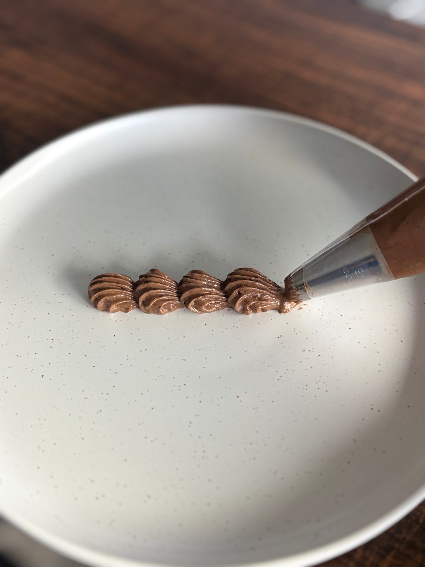 A close-up of a piping bag dispensing brown piped frosting or chocolate onto a white speckled plate, with a wooden table in the background.