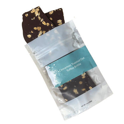 Oat brittle pieces spilling out of packaging labeled 'Cinnamon Toasted Oat Toffee Brittle' on a white background
