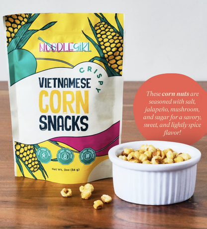 NoodleGirl Vietnamese Corn Snacks in a yellow and white stand-up pouch, shown next to a small white ramekin filled with corn nuts on a wooden surface. A red circular text bubble provides flavor and seasoning details.