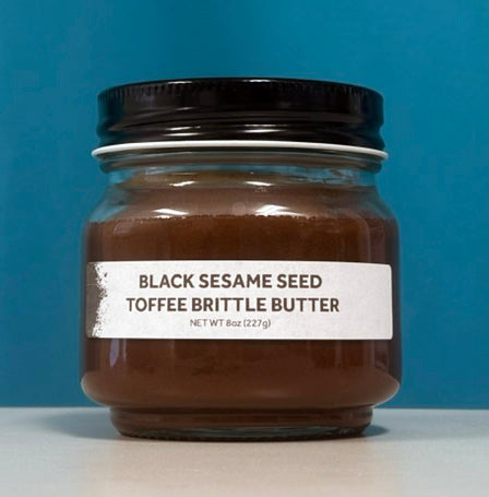 A jar of Black Sesame Seed Toffee Brittle Butter with a white label and black lid, set against a solid blue background.